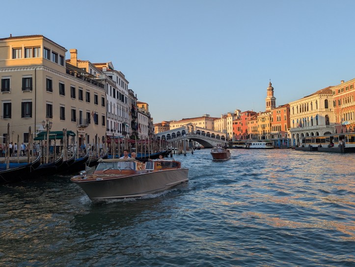 Evening view of Venice’s Grand Canal with water traffic, gondolas and historic palazzi lining the waterfront, leading towards the Rialto Bridge under a clear sky.