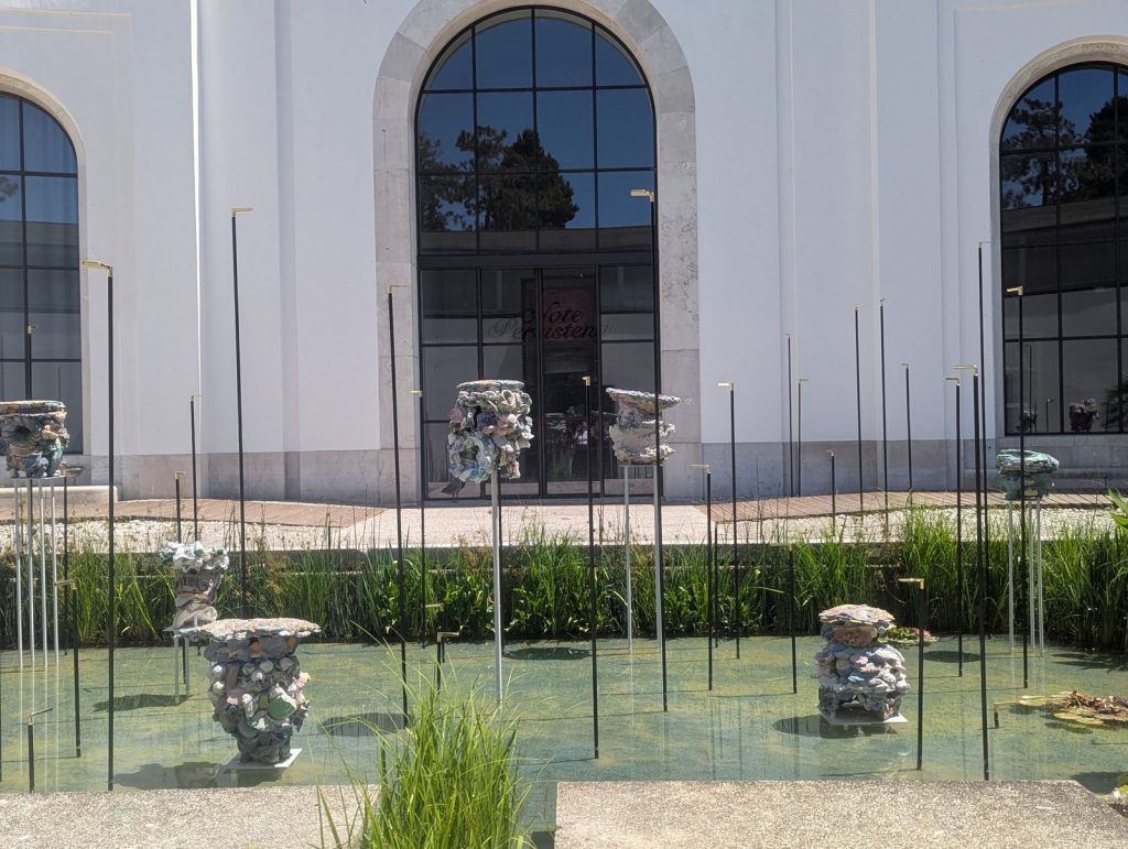 Outdoor sculptural installation set within a shallow reflecting pool beside a white pavilion building. Clusters of textured ceramic-like forms rise from the water among a grid of slender metal poles and aquatic planting.